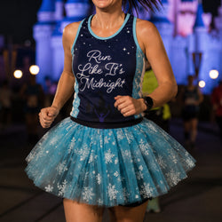 A woman running at night wearing a dark navy tank top with 'Run Like It's Midnight' in white script, paired with a light blue tutu skirt covered in white snowflakes. The background shows a blurred castle and event lights.