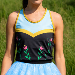An athletic-style tanktop worn by a model outdoors, featuring a light blue upper section with yellow trim, a black lower section with pink floral embroidery, and a light blue tutu skirt with white snowflake patterns.