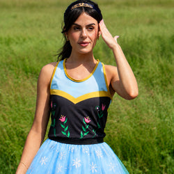 A woman wearing a Frozen-inspired tanktop with a black bodice featuring pink floral embroidery, a light blue yoke with yellow trim, and a light blue tulle tutu skirt with white snowflakes, set against a blurred green grassy background.