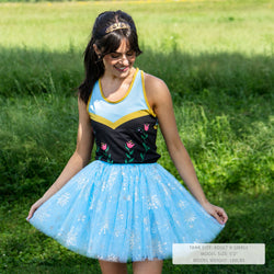 A woman standing in a grassy field wearing an athletic-style tanktop with a light blue and black body, yellow trim, and floral embroidery, paired with a light blue tutu skirt with white snowflake-like patterns.