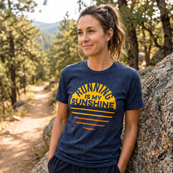 A woman wearing a navy blue heather graphic t-shirt with a yellow and orange sunburst design and the text 'RUNNING IS MY SUNSHINE' stands outdoors next to a large rock in a forest setting.