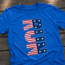 A royal blue short-sleeve t-shirt laid flat on a dark wooden surface, featuring a large vertical graphic spelling 'RUN' in an American flag pattern with red and white stripes and white stars.