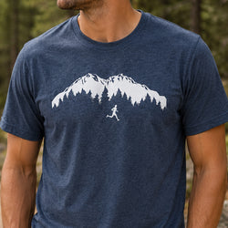 A man wearing a heather navy blue t-shirt with a white graphic of a mountain range, pine trees, and a running trail runner. The background is a blurred outdoor forest setting.