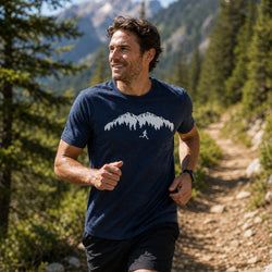 A man running on a forest trail wearing a navy blue t-shirt with a white mountain and runner graphic, paired with black shorts.