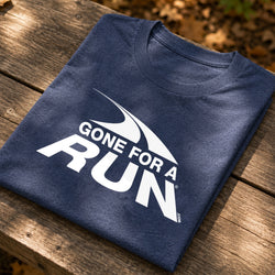A navy blue folded t-shirt with a white 'GONE FOR A RUN' graphic, displayed on a rustic wooden table with blurred autumn leaves in the background.
