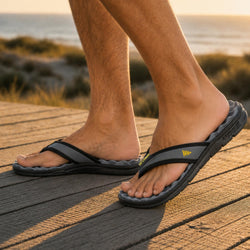 A close-up of a person's feet wearing black and gray slide sandals with a small yellow logo, standing on a wooden boardwalk with a beach and ocean in the background.
