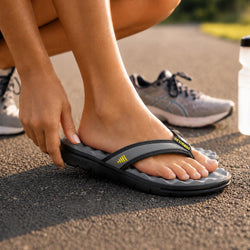 A close-up lifestyle shot of a person wearing a gray and black cushioned flip-flop sandal outdoors on asphalt, with their hand adjusting the strap. A gray running shoe is visible in the background.