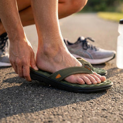 A close-up lifestyle shot of a person putting on an olive green and black water slide flip-flop sandal on asphalt, with a gray running shoe in the background.
