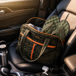A quilted olive green travel tote bag with orange trim sits on a car seat, filled with flip-flops. The bag has the brand name 'FLYTE' printed on the front.