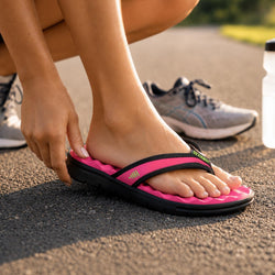 A close-up of a person wearing a bright pink and black orthopedic flip-flop sandal outdoors on asphalt, with their hand adjusting the strap. A gray running shoe is visible in the background.