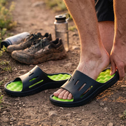 A person outdoors on dirt ground wearing black slide sandals with bright green textured footbeds, with hiking shoes and a water bottle in the background.