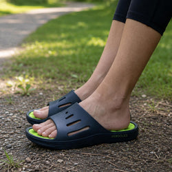 A person wearing black slide sandals with bright green insoles, standing on a dirt path outdoors with grass in the background. The brand name 'PRSOLES' is visible on the side of the sandal.