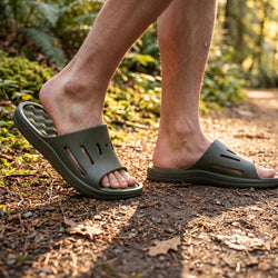 A person wearing dark olive green slide sandals walking on a forest path covered with pine needles and leaves.