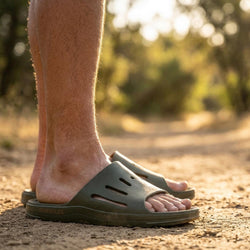 A close-up of a person's feet wearing dark olive green prsoles slide sandals on a dirt path outdoors with a blurred natural background.