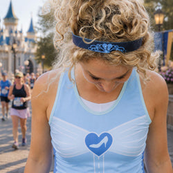 A woman in a light blue athletic tank top with a blue high-heel heart logo and a matching blue headband, looking down in a parade-like outdoor setting with a castle in the background.