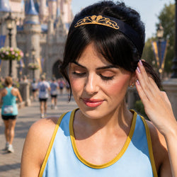 A woman outdoors wearing a black headband with an ornate gold tiara design, paired with a light blue top with gold trim. The background shows a blurred castle and park scene.
