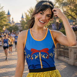 A woman wearing a blue and yellow princess-themed athletic top and yellow sequined tutu skirt, smiling in front of a blurred castle background with other runners visible.