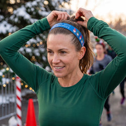 Woman in green long-sleeve shirt adjusting headband with festive gnome design, blurred Christmas trees and people in the background.
