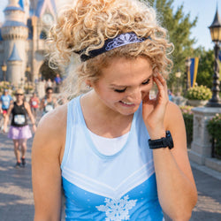 A woman with curly blonde hair wearing a light blue and white tanktop with a snowflake design, standing outdoors in front of a castle. She is smiling and touching her face, with a black smartwatch on her wrist.
