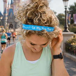 A woman with curly blonde hair wearing a light green tank top and a blue headband that reads 'Eat My Pixie Dust' in white script, standing outdoors in front of a castle in a park.