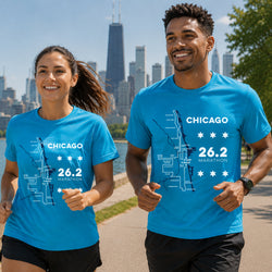 Two people jogging outdoors in matching bright blue Chicago 26.2 Marathon t-shirts, with a city skyline and water in the background.