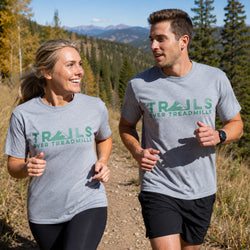 A man and a woman jogging outdoors on a trail, both wearing heather gray athletic t-shirts with the green text 'TRAILS OVER TREADMILLS' printed across the chest. The background shows a scenic mountain landscape with trees and a dirt path.