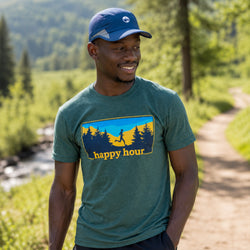 A man wearing a green graphic t-shirt and a blue baseball cap stands on a dirt path in a forest setting. The t-shirt features a graphic of a hiker with the text 'happy hour' in yellow.