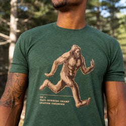 A man wearing a dark green heathered t-shirt with a graphic of a running Bigfoot and the text 'TRAIL RUNNING CHAMPION LOCATION UNKNOWN'.