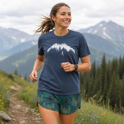A woman running outdoors on a mountain trail wearing a navy blue heathered t-shirt with a white mountain graphic and green patterned shorts, with a forest and mountains in the background.
