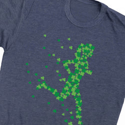 A close-up of a heather navy blue t-shirt featuring a graphic of a running figure composed of green shamrocks, with small shamrocks scattered around it.