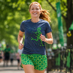 A woman running outdoors wearing a navy blue graphic t-shirt with a green shamrock pattern and matching green patterned athletic shorts.