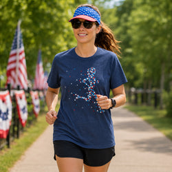 A woman jogging outdoors on a tree-lined path wearing a navy blue t-shirt with a patriotic red, white, and blue confetti graphic, a matching patriotic visor, and black athletic shorts.