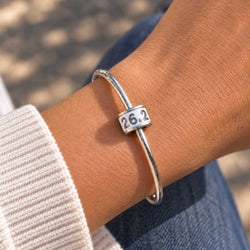 A close-up of a person's wrist wearing a polished silver bangle bracelet with a rectangular charm engraved with the number 26.2, representing the Colorado Trail Marathon.