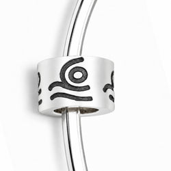 A silver-toned cylindrical charm bead with black engraved abstract symbols, shown threaded onto a matching silver bangle against a clean white background.