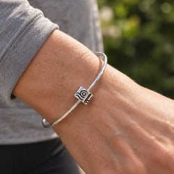 A sterling silver bangle bracelet with a small geometric charm featuring a spiral design, shown worn on a person's wrist.