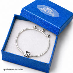 A silver-toned bangle bracelet with a textured hammered finish and a small decorative bead, displayed inside an open bright blue gift box with a white insert. The lid has a blue logo, and a small text note at the bottom says '*gift box not included'.