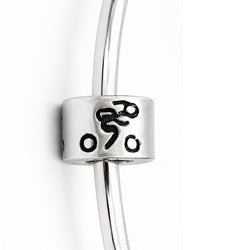 A close-up of a silver bangle bracelet featuring a cylindrical charm with black engraved symbols, including a bicycle.