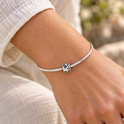 A close-up of a person's wrist wearing a thin silver bangle bracelet with a small silver cube charm engraved with black pips, set against a soft, blurred outdoor background.