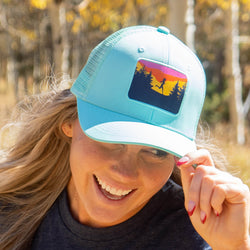 A woman outdoors wearing a light blue trucker hat with a colorful sunset landscape patch on the front panel.