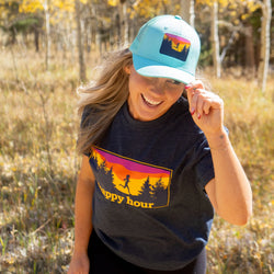 Woman wearing a light blue trucker hat and navy t-shirt featuring a sunset "happy hour" mountain runner design.