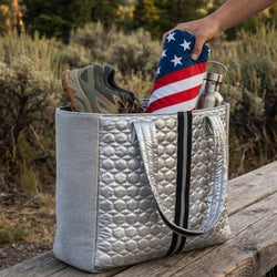 Silver tote bag with American flag towel, shoes, and water bottle on a wooden surface outdoors.