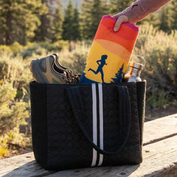Black tote bag with  running shoes, colorful towel, and bottle on a wooden surface with a natural background.