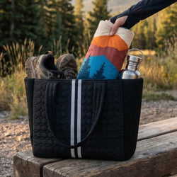 Black tote bag with hiking gear including shoes, a colorful towel, and a water bottle, set against a natural background.