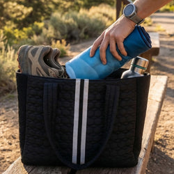 Black bag with a blue towel, shoes, and a silver flask on a wooden surface outdoors.