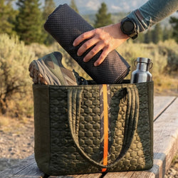 Person holding a rolled-up towel over a green quilted bag with hiking shoes and a water bottle in a natural setting.