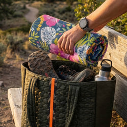 Person holding a floral-patterned towel over a backpack with hiking shoes and a water bottle in a natural setting.