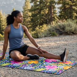 Woman stretching on a colorful mat outdoors with trees and mountains in the background