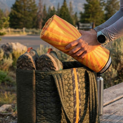 Person holding a orange and yellow towel next to a quilted bag outdoors.