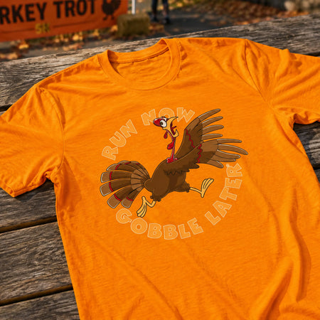 An orange short-sleeve t-shirt laid flat on a weathered wooden table, featuring a cartoon turkey graphic with the text 'RUN NOW GOBBLE LATER'.