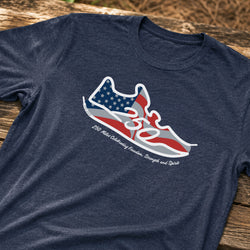 A navy blue heather t-shirt laid flat on a rustic wooden surface, featuring a graphic of a sneaker filled with an American flag pattern and the text '250 Miles Celebrating Freedom, Strength and Spirit'.